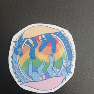 May include: A circular sticker with two blue dragon illustrations, set against a rainbow background. The dragons have yellow markings and are in a playful pose. The sticker is edged with a white border.