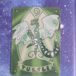 May include: A green card with a cartoon dragon holding a swirl. The dragon has white spots and wings. The word "TURTLE" is written on a banner at the bottom of the card. The card has a green border and a light green background.