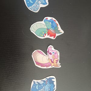 May include: Four colourful dragon stickers. The stickers feature various dragon designs in shades of blue, green, pink, and red. The dragons have unique patterns and expressions, with a cartoonish style. The stickers are on a black surface.