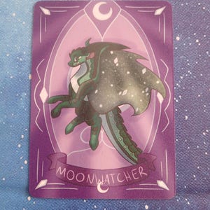 May include: A purple card with a dragon illustration in green and white. The dragon has speckled wings and is set against a decorative background with a crescent moon. The text "MOONWATCHER" is displayed on a banner across the bottom of the card.