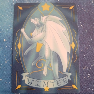 May include: A rectangular card featuring a light blue dragon with large white wings, set against a teal and navy background. The word "WINTER" is displayed on a banner. Gold stars and geometric shapes frame the dragon.