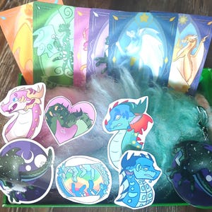 May include: A green box containing dragon-themed stickers and cards. The stickers depict dragons in pink, blue, and green. The cards feature dragon illustrations with stained-glass backgrounds. The box also holds teal-coloured fluff.
