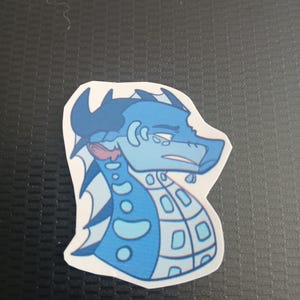May include: A sticker depicting a blue dragon with a melancholic expression. The dragon has light and dark blue scales, a pink throat, and a white outline. The sticker is on a black surface.
