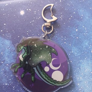 May include: A clear acrylic keyring featuring a green dragon with a starry wing design. The dragon is set against a purple background with crescent moon and star details. The keyring has a silver-coloured clasp.