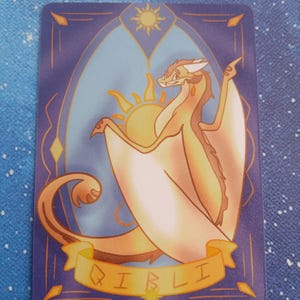 May include: A trading card with a dragon illustration. The dragon is tan and brown with white wings, set against a blue background with a sun design. The name "QIBLI" is written on a banner.