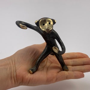 Puede incluir: Una pequeña figura de mono de bronce con detalles dorados. El mono está de pie sobre sus patas traseras con un brazo levantado y el otro doblado en el codo. El mono tiene una cara estilizada con ojos dorados y una boca dorada.