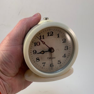 Pode incluir: Um despertador vintage branco com números e ponteiros pretos. O mostrador do relógio diz "Celann" e tem um fundo branco. O relógio está sendo segurado na mão.