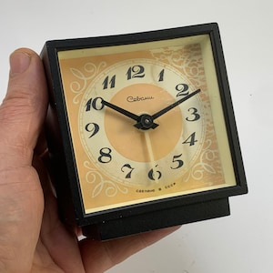 Pode incluir: Um relógio quadrado preto com um mostrador de cor creme e ponteiros pretos. O mostrador do relógio tem algarismos romanos pretos e a palavra "Сальск" impressa em preto. O mostrador do relógio também tem um padrão decorativo ao redor da borda e o texto "Сделано в СССР" impresso na parte inferior.