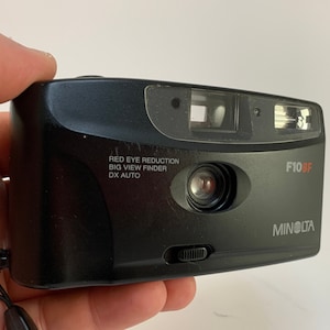 May include: A black Minolta F10BF point-and-shoot camera with a red eye reduction feature, a big view finder, and DX auto. The camera has a lens cover and a flash.