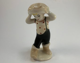 May include: A porcelain figurine of a child wearing a white hat, a white shirt with red polka dots, blue overalls, and white boots. The child is holding the hat with both hands and looking down.