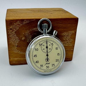 May include: A vintage stopwatch with a cream-colored face, silver casing, and a ring at the top. The face displays numbers and markings for measuring time. It is set against a wooden block background.