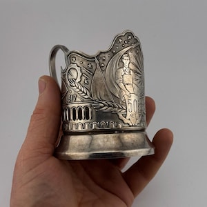 May include: Silver-toned metal cup holder with a handle. The cup holder is decorated with relief images of a man, a flag, a star, and the number "50". The year "1917" is also visible.