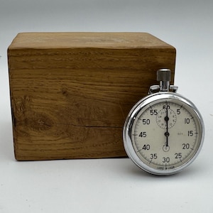 Peut inclure: Un chronomètre analogique argenté avec un cadran blanc, des chiffres noirs et une couronne de remontage sur le dessus, placé devant un bloc rectangulaire en bois brun naturel avec un grain visible et une fissure horizontale sur la face avant.