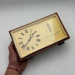 Puede incluir: Una combinación vintage de reloj y barómetro con marco de madera. La esfera del reloj tiene números romanos y manecillas negras. El barómetro muestra una escala de temperatura. El reloj es rectangular y tiene un borde plateado.