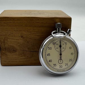 May include: A silver-toned analog stopwatch with a cream-colored face, black numerals, and a small sub-dial. The stopwatch features a winding crown and a start-stop button at the top, with the text MADE IN USSR printed at the bottom of the dial.