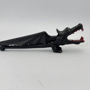 May include: A black dragon-shaped bottle opener with red accents on the mouth and teeth. The dragon has detailed wings and a long body. The opener is made of metal and is designed to open bottles.