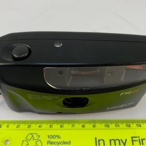 May include: A black Minolta F10BF point and shoot camera with a green body and a red eye reduction feature. The camera has a built-in flash and a viewfinder.
