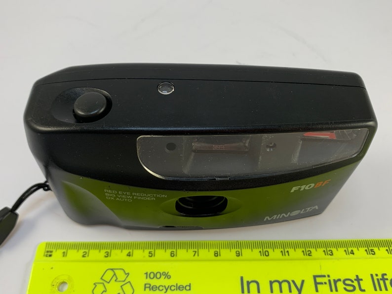 May include: A black Minolta F10BF point and shoot camera with a green body and a red eye reduction feature. The camera has a built-in flash and a viewfinder.