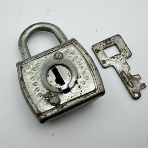 Puede incluir: Un candado plateado vintage con su llave. El candado tiene un cuerpo cuadrado con detalles decorativos y un asa curva. La llave tiene una forma rectangular simple con un diseño de ojo de cerradura.