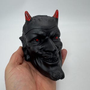 May include: A black devil head figurine with red horns and red eyes. The figurine is made of a hard material and has a detailed face.