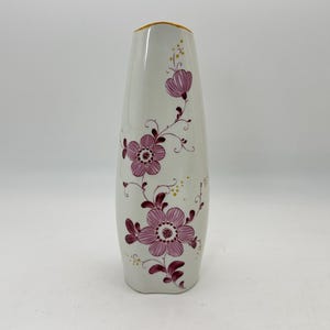 May include: A white ceramic vase with a slightly flared top and a gold rim. The vase is decorated with a floral pattern in shades of pink and burgundy. The flowers are arranged in a flowing, vine-like design.