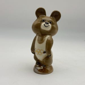 May include: A brown ceramic bear figurine wearing a white and gold Olympic uniform. The bear has a smiling face and is standing on a brown base. The Olympic rings are visible on the uniform.