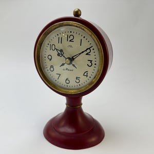 May include: A red tabletop clock with a white face and black hands. The clock has a gold rim and a base with a rounded top. The clock face reads "Maxx".
