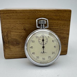 Puede incluir: Un cronómetro vintage con caja plateada y esfera color crema, que muestra números y marcas. El cronómetro está sobre un fondo de bloque de madera. La esfera indica "Azam" y "СДЕЛАНО В СССР".