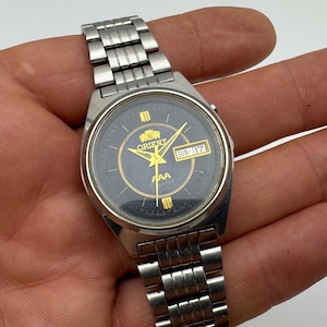 Könnte beinhalten: Silberfarbene Orient-Armbanduhr mit schwarzem Zifferblatt und goldenen Akzenten. Die Uhr zeigt Tag und Datum, mit "SON-17" sichtbar. Sie hat ein silbernes Metallarmband.