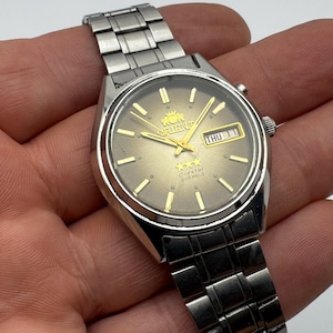 Può includere: Un orologio Orient Crystal argentato con quadrante e lancette dorate. Il quadrante mostra il giorno e la data, con la scritta "Orient" in alto. Ha un cinturino in metallo argentato.