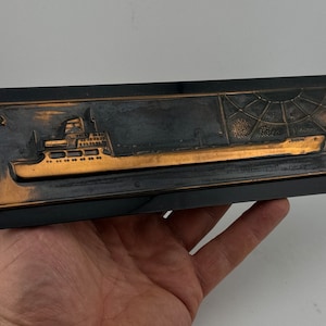 May include: A black framed plaque featuring a copper-coloured relief of a ship. The ship is detailed with windows and a deck. The background is a dark copper colour with a spiderweb design.