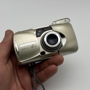 May include: A silver Olympus µ [mju:]-III 120 camera. The compact camera features a zoom lens, a built-in flash, and the Olympus logo. The lens has the text "MULTI-AF ZOOM 38-120mm".