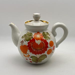 May include: White ceramic teapot with a floral design. The teapot features a large red and orange flower with green leaves, and orange accents. The lid and handle are white, and the rim is gold.