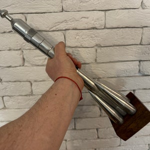 Può includere: Una scultura di razzo argentata con una base in legno. Il razzo è composto da più sezioni che sono collegate tra loro. La scultura è tenuta da una mano.