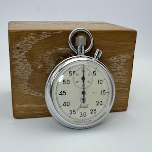 Könnte beinhalten: Eine Vintage-Stoppuhr mit silbernem Gehäuse und cremefarbenem Zifferblatt, das Zahlen und Markierungen zur Zeitmessung anzeigt. Sie liegt vor einem Holzblock. Der Text "Azam" und "Made in USSR" sind sichtbar.