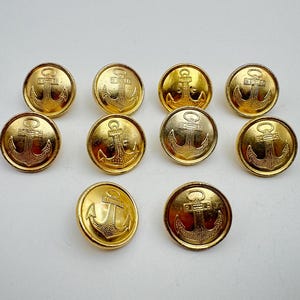 Könnte beinhalten: Zehn goldfarbene Knöpfe mit Anker-Design. Die Knöpfe sind rund und haben eine glänzende Oberfläche. Der Anker befindet sich in der Mitte jedes Knopfes.
