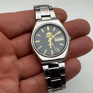 May include: A silver-toned Orient wristwatch featuring a round black dial with gold-colored hour markers and hands. The watch includes a day-date window at the three o'clock position displaying the text SUN 27 and a stainless steel link bracelet.