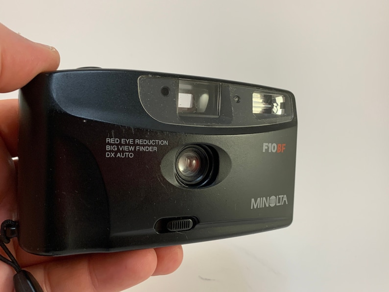 May include: A black Minolta F10BF point-and-shoot camera with a red eye reduction feature, a big view finder, and DX auto. The camera has a lens cover and a flash.