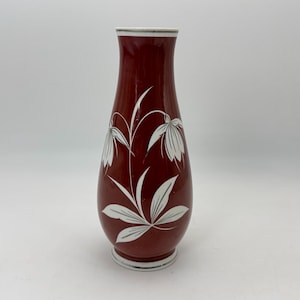 May include: A maroon ceramic vase with a white floral design. The vase has a flared top and a round base with a white rim. The floral design features stylized white flowers and leaves.