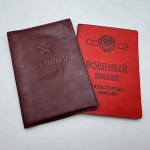Puede incluir: Dos libretas de identificación militar soviéticas rectangulares. Una es de color granate oscuro con una estrella en relieve y el texto VOENNYY BILET. La otra es de color rojo brillante con el texto impreso en negro SSSR, VOENNYY BILET, MINISTERSTVO OBORONY y un escudo.