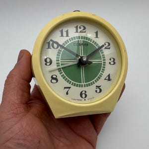 Può includere: Una sveglia vintage color crema con quadrante rotondo. Il quadrante presenta numeri neri, lancette nere e un centro verde. L'orologio è contrassegnato con la parola "SEVANI" e il testo "Made in USSR".