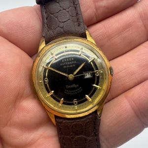 Puede incluir: Un reloj de pulsera dorado con una esfera negra y una correa de cuero marrón. La esfera del reloj tiene marcas blancas y el texto "Anker Automatic 30 Rubis" y "Tissot Waterproof Ander 5 ATM".