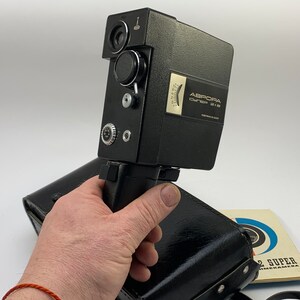 May include: A black Aurora Super 8 film camera with a leather carrying case and instruction manual. The camera has a zoom lens and a built-in microphone.