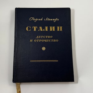 Può includere: Un libro rilegato in blu con lettere dorate intitolato "Stalin: Infanzia e adolescenza" di Georgi Leonidze.