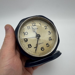 Può includere: Una sveglia blu con quadrante bianco e lancette nere. L'orologio ha una cassa in plastica nera e un quadrante bianco con numeri neri. L'orologio è stato realizzato in URSS e riporta la scritta "Севани" sul quadrante.