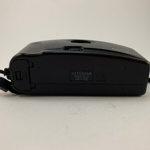 May include: Black Minolta camera with a black strap. The camera has a serial number 317750850 and the text "Minolta Co., Ltd. Osaka, Japan Made in China" printed on the bottom.