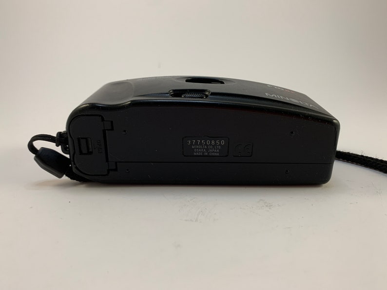 May include: Black Minolta camera with a black strap. The camera has a serial number 317750850 and the text "Minolta Co., Ltd. Osaka, Japan Made in China" printed on the bottom.