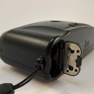 May include: A black compact camera with a silver battery compartment door open. The camera has a black strap attached to it.