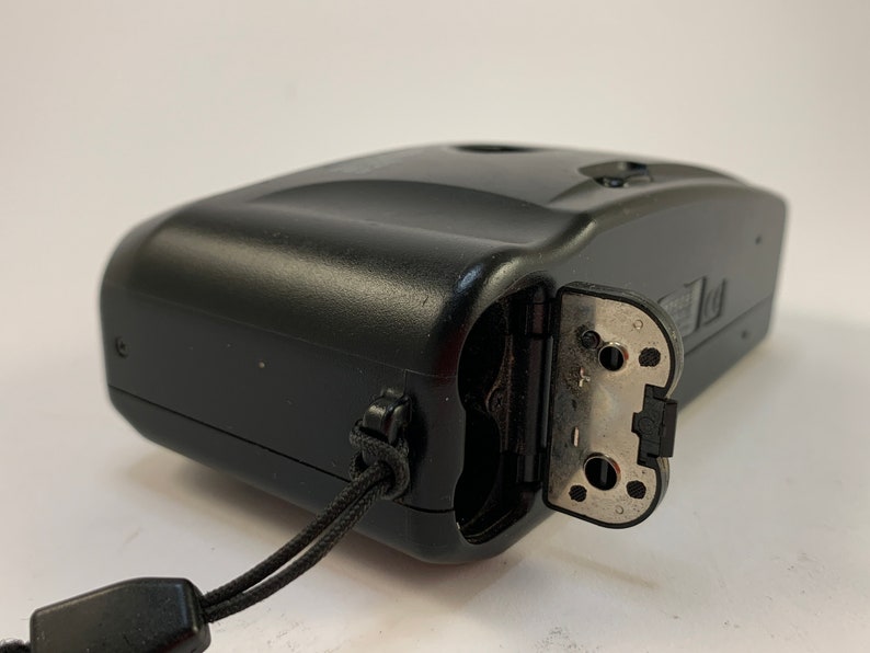 May include: A black compact camera with a silver battery compartment door open. The camera has a black strap attached to it.