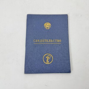 Peut inclure: Un livret bleu avec des lettres et des emblèmes dorés. Le texte "СВИДЕТЕЛЬСТВО" est centré sur la couverture. Le livret a un aspect texturé et repose sur une surface blanche.